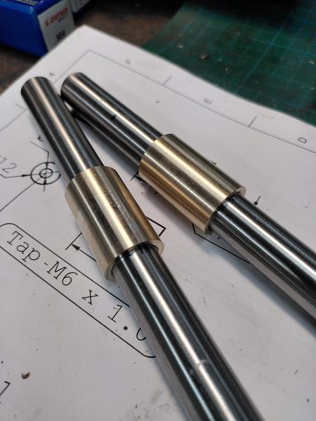 Two polished stel bars laying on a technical drawing. Each bar has a
bronze sleeve mounted on it. The only visible part of the drawing says
"Tap M6 x 1.0".