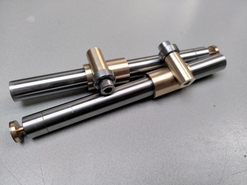 Two steel bars laying on a grey table. Each steel bar has a bronze
sleeve. Each bronze sleeve has a small brass cylinder sitting
perpendicular across the top. Each brass cylinder has a wide thick steel
washer attached with a stainless steel hex cap head screw. In the end of
each steel bar is a bronze screw.