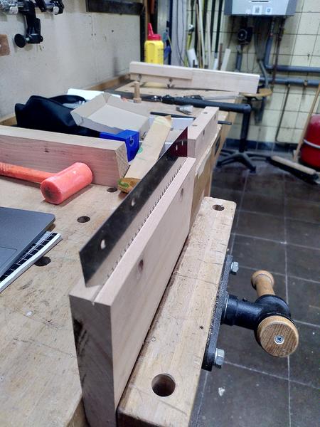 A piece of beech (fagus sylvatica) held in the vice on a workbench. A
unhandled saw blade is making a groove in the top of the wood. The
workbench beyond is cluttered with tools and materials.